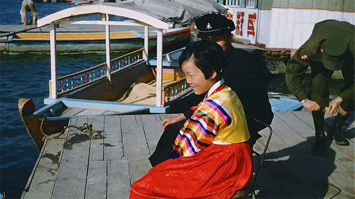 Một cô gái trẻ ngồi dọc sông Hàn mặc hanbok sặc sỡ, phong cách đặc trưng của các cô gái trẻ trong thời kỳ này. Bên cạnh cô là một học sinh, mặc đồng phục đen và đội mũ lưỡi trai.