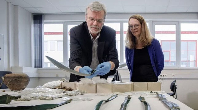 Các nhà khảo cổ học Detlef Jantzen (trái) và Bettina Martin, Bộ trưởng Bộ Khoa học và Văn hóa, xem những phát hiện khảo cổ mới nhất từ Đức, bao gồm những thanh kiếm thời kỳ đồ đồng và hàng nghìn đồng bạc.(Ảnh: Markus Scholz/dpa via Alamy Live News)